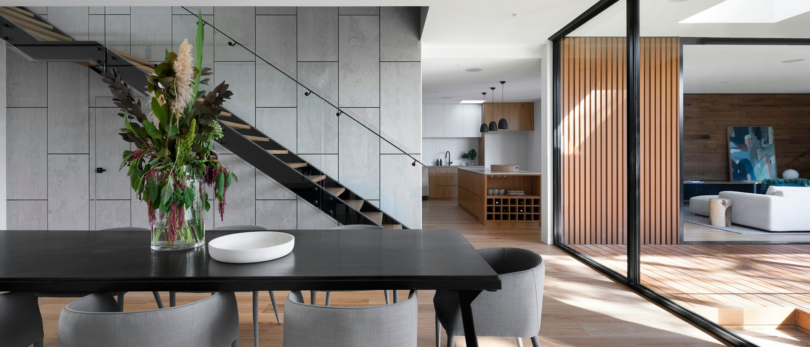 Dining table beneath a floating stair, with the kitchen and a timber-slatted garden door beyond